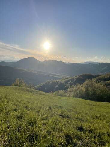 Um sol brilhante ilumina as colinas verdes e onduladas e as montanhas distantes, banhando uma paisagem tranquila com céu azul, nuvens esparsas e campos verdejantes lá embaixo.