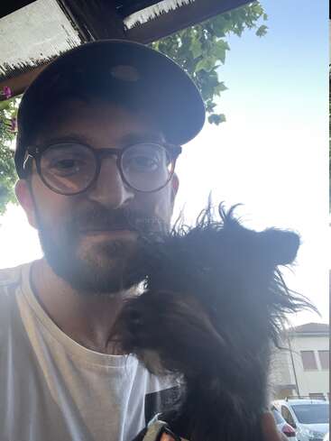 Um homem de óculos e boné tira uma selfie com seu cachorrinho preto e desgrenhado lá fora. No fundo, dá pra ver árvores, o céu e prédios.
