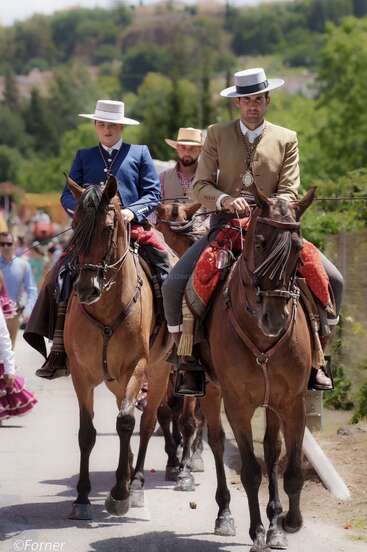 Deux cavaliers en tenue traditionnelle espagnole, portant des chapeaux, montent des chevaux bruns lors d'un défilé culturel. Un autre cavalier suit derrière. L'arrière-plan est constitué d'une végétation luxuriante et de personnes.