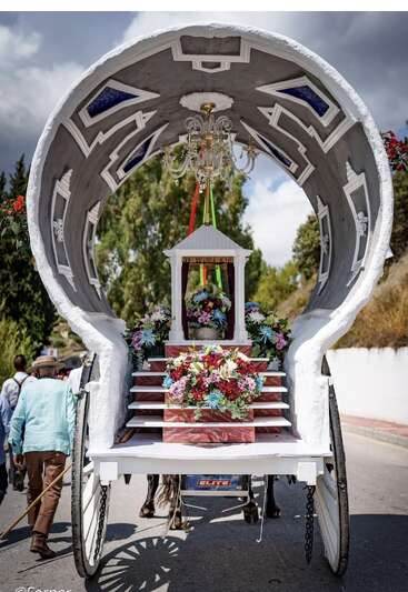 Un chariot magnifiquement décoré de compositions florales, d'un chandelier et d'une châsse est tiré lors d'une procession. Des gens marchent à côté sous un ciel nuageux.