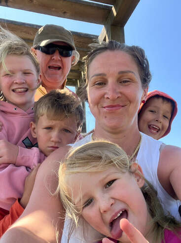 Un groupe de cinq personnes heureuses, comprenant des adultes et des enfants, prend un selfie joyeux en plein air sous une structure en bois, profitant ensemble d'une journée ensoleillée.