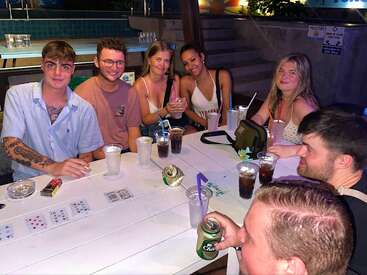 Seven friends sit around a white table outdoors at night, enjoying drinks and playing cards. The atmosphere is relaxed, cheerful, and illuminated by soft lighting.