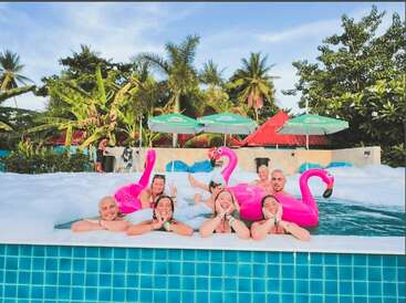 A group of friends enjoy a fun pool party with foam, colorful inflatable flamingos, and tropical palm trees in the background, laughing under the sunny sky.