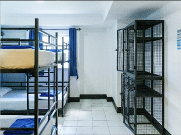 This image shows a clean hostel dormitory room with metal bunk beds, blue linens, secure black lockers, white tiled floors, and a bright window with blue curtains.