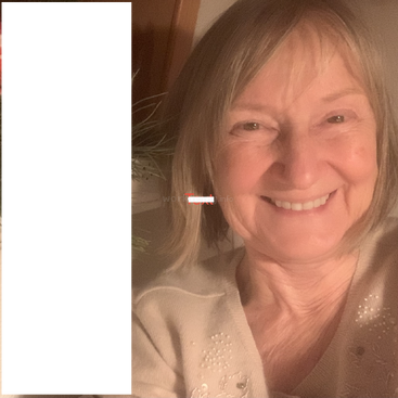 Uma mulher sorridente, de cabelo curto e claro, usa um suéter de cor clara decorado com miçangas. Ela posa dentro de casa, perto de uma planta. O lado esquerdo está escurecido.