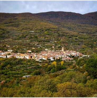Das Bild zeigt eine kleine Stadt in den Bergen, umgeben von üppigem Grün und Bäumen, mit einem bewölkten Himmel darüber.