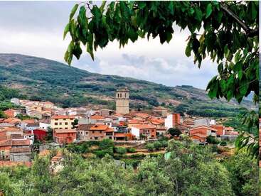 Das Bild zeigt eine kleine Stadt in den Bergen mit einem markanten Kirchturm und einer Ansammlung von Gebäuden mit Terrakotta-Dächern, umgeben von üppigem Grün.