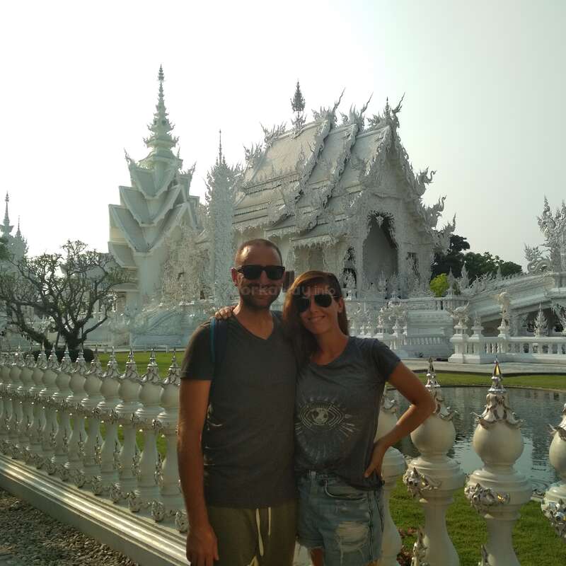 Das Bild zeigt einen Mann und eine Frau, die vor einem weißen Tempel posieren, mit einem verzierten Geländer und einem ruhigen Teich im Vordergrund vor einem strahlenden Himmel.