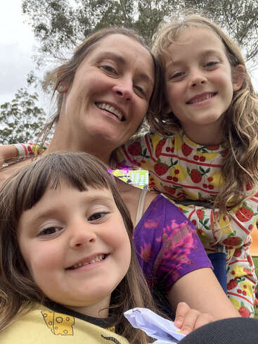 Eine glückliche Frau und zwei lächelnde junge Mädchen posieren eng zusammen im Freien. Die Kinder tragen bunte Kleidung mit Obst- und Käseaufdrucken. Der Hintergrund ist mit Bäumen gefüllt.