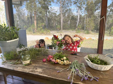 Zwei lächelnde Kinder posieren hinter einem Holztisch, der mit Blumen, Grünzeug und Gartengeräten gefüllt ist, während sie an einem sonnigen Tag von Natur und Bäumen umgeben sind.