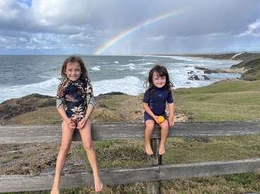 Zwei lächelnde Kinder sitzen auf einem Holzzaun über einer grasbewachsenen Klippe und halten Früchte in der Hand, während sich hinter ihnen das Meer, die Wellen und ein leuchtender Regenbogen wölben.