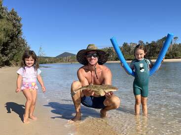 Ein Mann mit Hut und Sonnenbrille kniet an einem Sandstrand und hält einen Fisch. Er lächelt mit zwei jungen Mädchen, eines im Badeanzug, eines mit Poolnudeln.
