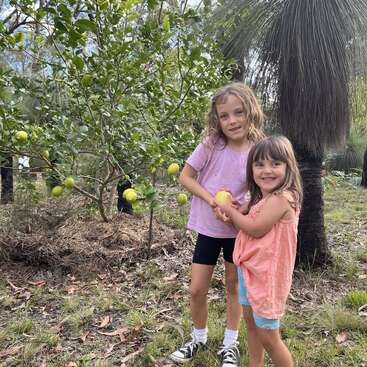 Two young girls stand outside by a lemon tree, smiling and holding a lemon together. The scene is natural, happy, and surrounded by greenery and trees.