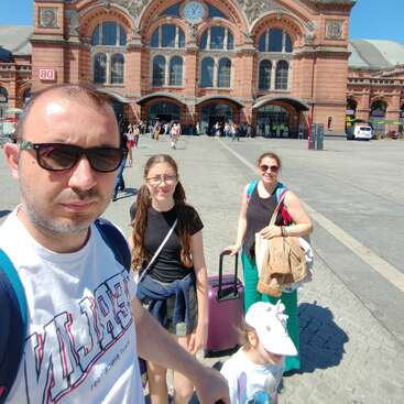 Eine vierköpfige Familie steht an einem sonnigen Tag vor einem historischen Bahnhof, mit Gepäck und in legerer Kleidung, bereit für Reisen und Abenteuer.