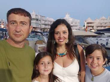 Eine lächelnde vierköpfige Familie posiert an einem Yachthafen mit Yachten und weißen Gebäuden im Hintergrund und genießt einen gemeinsamen Abend am Wasser.