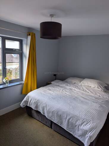 This bedroom features a neatly made double bed with white linens, light gray walls, a window with bright yellow curtains, and a small vase of flowers.