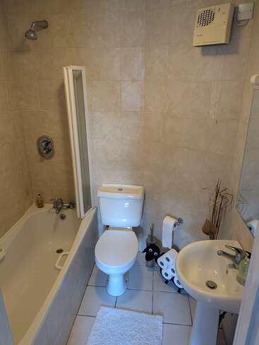 This is a compact, clean bathroom with beige tiles, bathtub, shower, toilet, pedestal sink, white mat, and whimsical sheep-shaped toilet paper holder beside the sink.