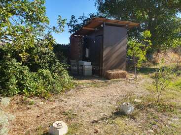 Une petite dépendance extérieure avec un toit en tôle ondulée se trouve parmi les arbres et les buissons. Une balle de foin, un chemin de pierre et un lavabo se trouvent à proximité. Journée ensoleillée.