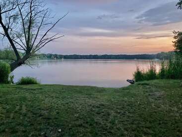 Das Bild zeigt eine ruhige Seeszene bei Sonnenuntergang, mit einem grasbewachsenen Ufer, einem blattlosen Baum und einem Boot, mit einem leuchtenden Himmel und fernen Bäumen.
