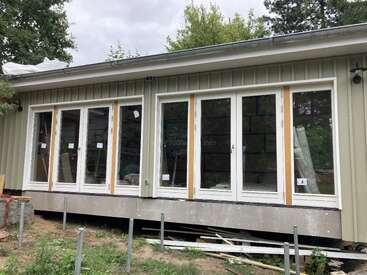 Dieses Bild zeigt ein im Bau befindliches Haus mit großen Glastüren und Fenstern. Das Äußere ist hellgrün, umgeben von Bäumen, und darunter sind Baumaterialien zu sehen.