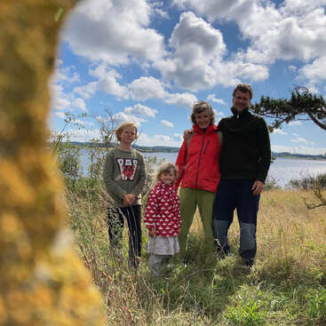 L'image représente une famille de quatre personnes debout dans un champ herbeux, avec une étendue d'eau sereine et un ciel bleu avec des nuages blancs en arrière-plan.