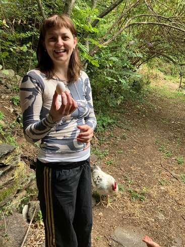 Eine lächelnde Frau steht draußen in einer grünen Gegend und hat ein kleines Ei in der Hand. Ein weißes Huhn sitzt neben ihr auf dem erdigen Boden und macht die ländliche Szene noch schöner.