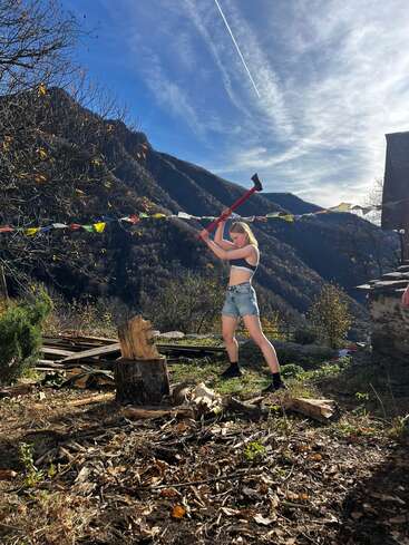 Eine Frau in Shorts und einem bauchfreien Top schwingt eine Axt, um draußen Holz zu spalten, umgeben von Bergen, herbstlichen Bäumen, Gebetsfahnen und strahlendem Sonnenschein.