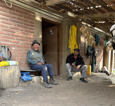 Two men, one elderly and one younger, sit and chat outside a rustic brick house. Corn hangs drying. They look relaxed and happy, enjoying each other’s company.
