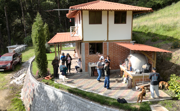 A group of people and several dogs gather outside a two-story house with a red roof, surrounded by greenery, preparing food near an outdoor oven.