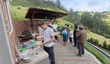 A young man prepares food outdoors at a rustic kitchen, while a small group of people socialize nearby, set against a scenic, green, mountainous landscape.
