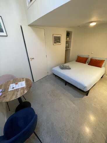 Dieses gemütliche, minimalistische Zimmer verfügt über ein Doppelbett mit orangefarbenen Kissen, ordentlich gefaltete Handtücher, einen kleinen runden Tisch mit zwei Stühlen, Betonboden und eine einfache Einrichtung.