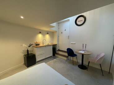 Dieses gemütliche, moderne Studio verfügt über eine Küchenzeile, einen runden Esstisch mit zwei Stühlen, eine Uhr, eine minimalistische Einrichtung, eine Treppe zu einer Tür und neutral getönte Wände.