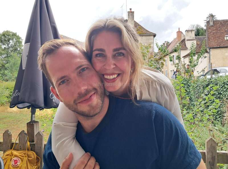 Ein lächelndes Paar posiert eng zusammen im Freien, wobei die Frau den Mann von hinten umarmt. Sie scheinen glücklich zu sein und genießen einen schönen Tag in einem malerischen Dorf.