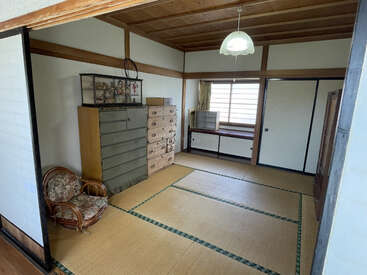 Se trata de una habitación tradicional japonesa con tatamis, puertas correderas, muebles de madera, una vitrina de cristal, una silla de ratán, una ventana y una lámpara de techo colgante.