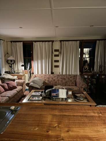 Ein gemütliches Wohnzimmer mit geblümten Sofas, einem mit Büchern gefüllten Couchtisch aus Holz und weißen Vorhängen. Die warme Beleuchtung schafft eine gemütliche, einladende Atmosphäre, die man von der Küche aus sieht.