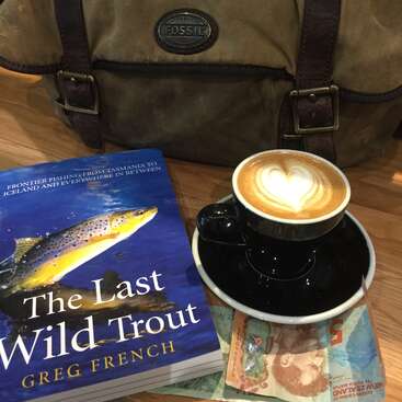 Ein Cappuccino mit herzförmigem Milchkaffee steht auf einer Untertasse, neben einem Buch mit dem Titel "Die letzte wilde Forelle", einer neuseeländischen Währung und einer Fossil-Tasche.