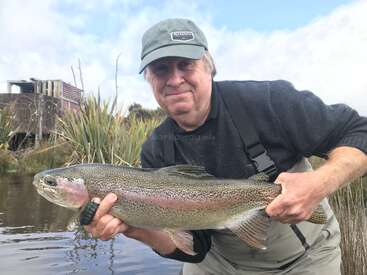 Ein Mann in Angelausrüstung hält stolz eine große Regenbogenforelle in der Nähe eines Flussufers, mit hohen Gräsern, Wasser und einer alten Holzkonstruktion im Hintergrund.