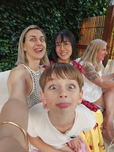 Quatre personnes posent pour un selfie ludique ; un enfant et une femme tirent la langue, tandis que deux femmes sourient, assises en plein air sur un fond de feuilles.