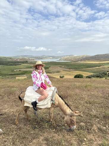 Une femme portant un chapeau et une chemise colorée monte un âne dans un paysage ouvert avec des collines ondulantes, des champs verts et un lac lointain sous les nuages.