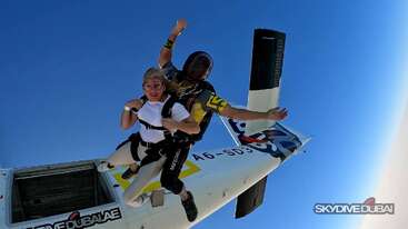 Un couple de parachutistes en tandem saute d'un avion de Skydive Dubai dans un ciel bleu limpide. La femme semble excitée, tandis que l'instructeur guide la descente.