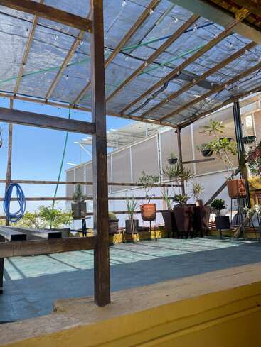 A rooftop terrace with a transparent roof, wooden frame, and potted plants. Bright natural light, light blue sky, and cozy outdoor setting create a peaceful atmosphere.