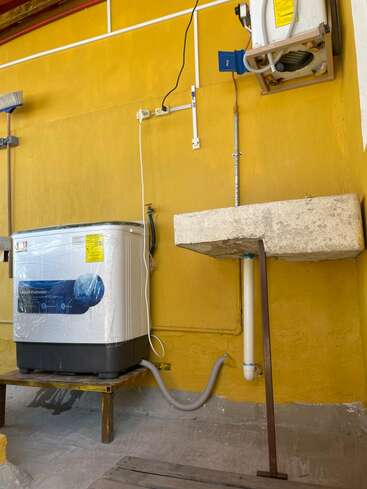 The image shows a yellow-walled laundry area with a washing machine, a concrete sink, a mounted faucet, a broom, pipes, electrical outlets, and an air conditioning unit.