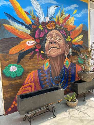 A vibrant mural depicts an elderly woman wearing colorful feathers and flowers in her headdress. She gazes upward, surrounded by mountains, plants, and a bright blue sky.