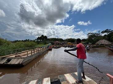 Un homme en chemise rouge se tient sur un quai en bois au bord d'une rivière, dirigeant un radeau à l'aide d'une perche. Il est entouré de bateaux colorés et d'une végétation luxuriante.