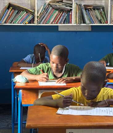 Trois garçons sont assis à des pupitres en bois, concentrés sur leurs copies d'examen. Des étagères de livres tapissent le mur au-dessus d'eux, créant une atmosphère studieuse et déterminée dans la salle de classe.