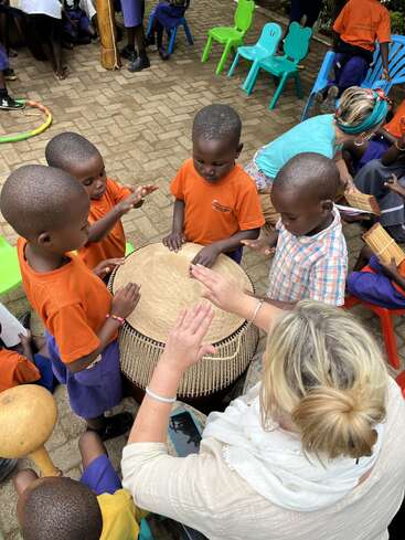 Plusieurs enfants et un adulte se rassemblent joyeusement autour d'un grand tambour en plein air, apprenant et jouant de la musique ensemble. Des chaises et des vêtements colorés ajoutent de l'éclat à la scène.