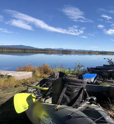 Un lac calme reflète un ciel bleu clair parsemé de nuages vaporeux. Des kayaks et des pagaies reposent sur la rive herbeuse, entourée de forêts et de montagnes lointaines.