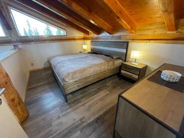 Cette chambre mansardée confortable dispose d'un plafond en bois avec poutres apparentes, de grandes fenêtres, d'un lit confortable, de tables de chevet modernes, de deux lampes et d'une petite commode.