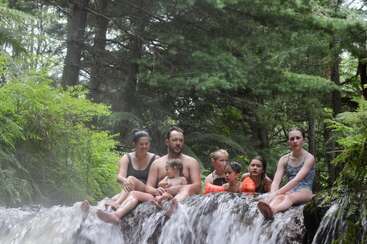 Uma família de sete pessoas senta-se nas rochas acima de uma cachoeira, cercada por uma floresta verdejante, curtindo uma aventura refrescante ao ar livre, todos de roupa de banho.