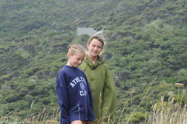Duas meninas estão ao ar livre, uma com um moletom azul do “Clube Atlético” e a outra com uma jaqueta verde, cercadas por grama alta e colinas verdes exuberantes.